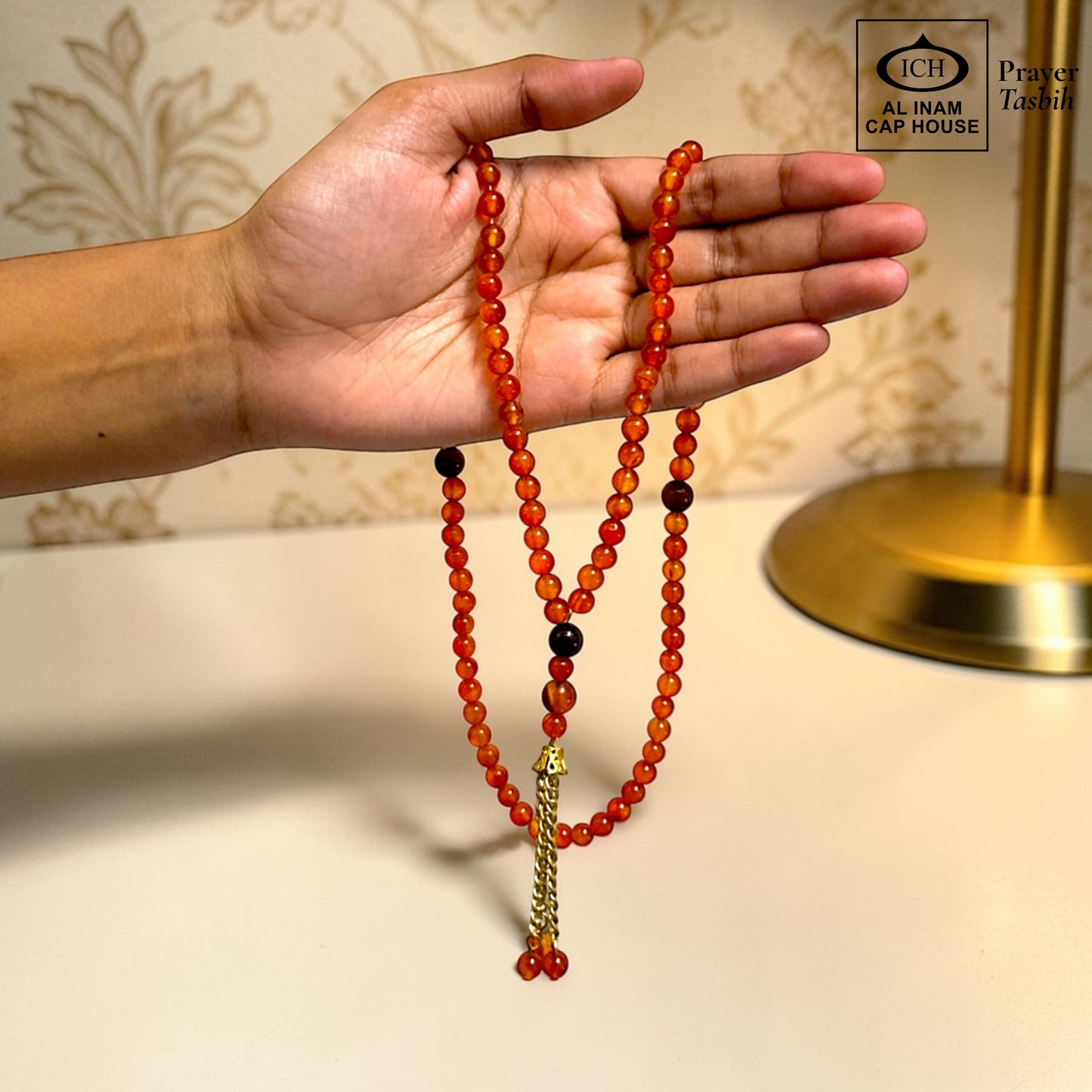 AL INAM CAP HOUSE amber and brown crystal tasbih with gold tassel, held in a hand.